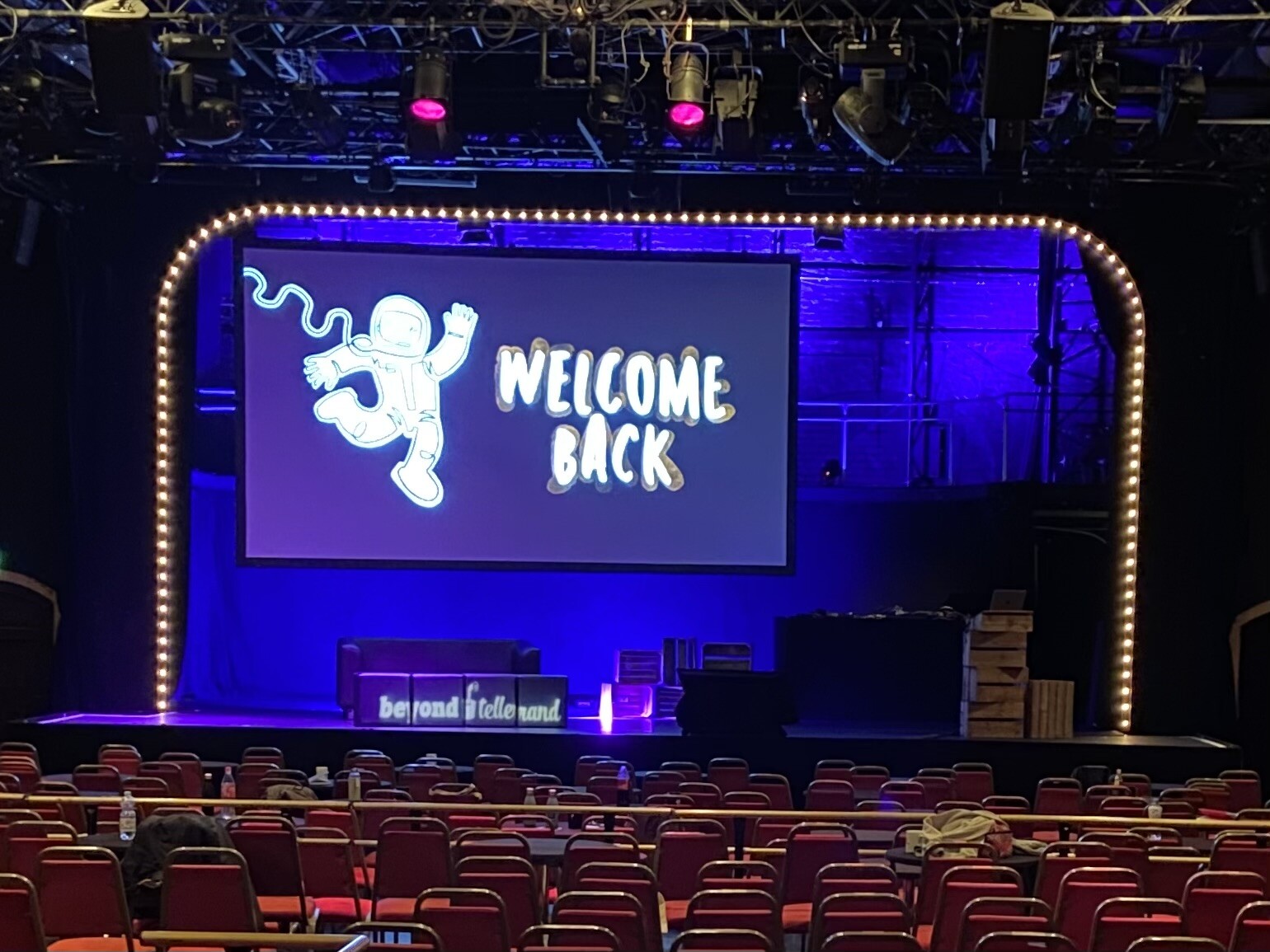 A stage in an empty thater with a big screen showing the words “welcome back“.