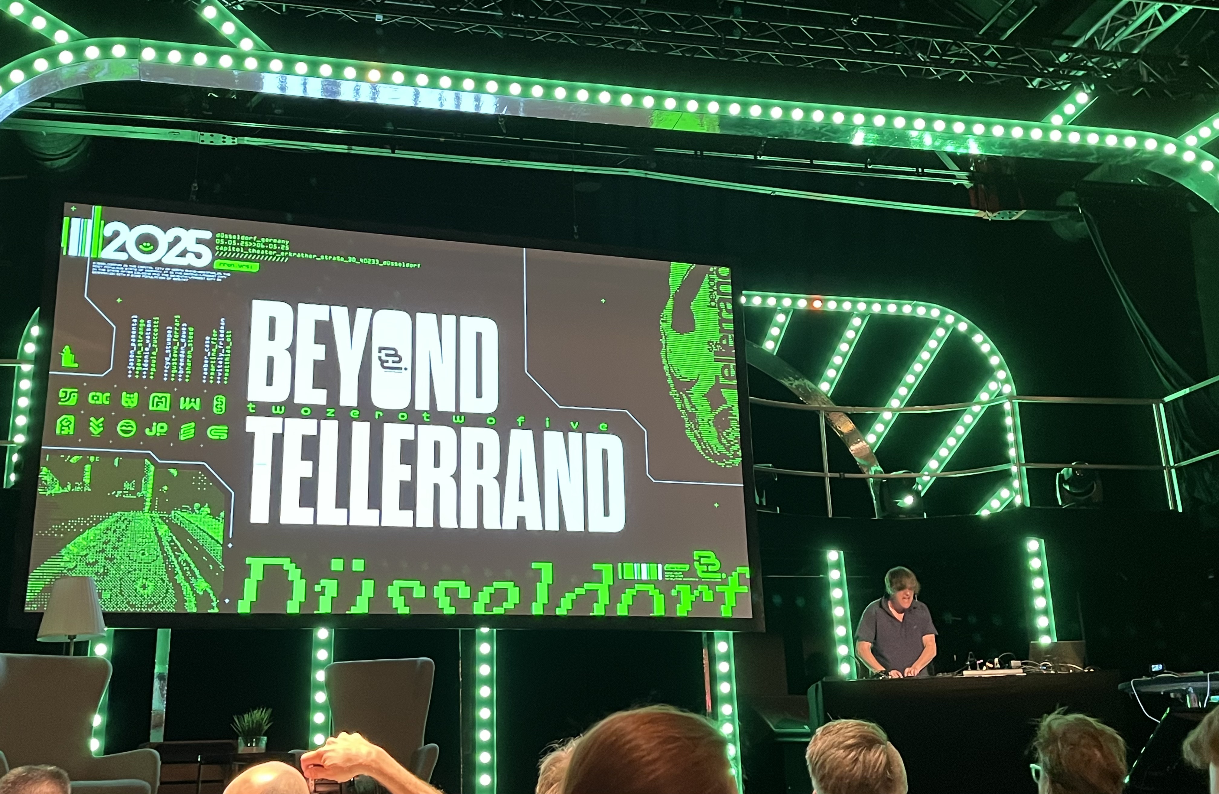 A green-lit stage with a large screen on which “beyond tellerrand” is written in white and green colors. On the right-hand side, a DJ can be seen at a console.
